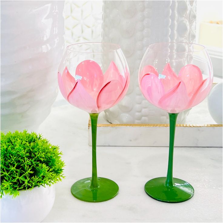 two wine glasses with pink flowers in them sitting on a counter top next to a potted plant