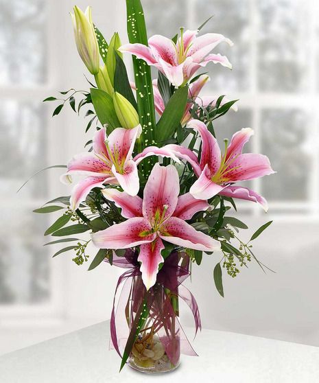 a vase filled with pink lilies and greenery on a white table next to a window