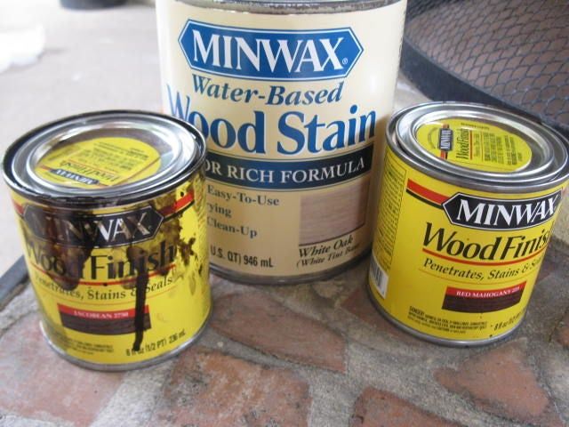 three cans of wood stain sitting next to each other on a brick patio area with a metal grill in the background