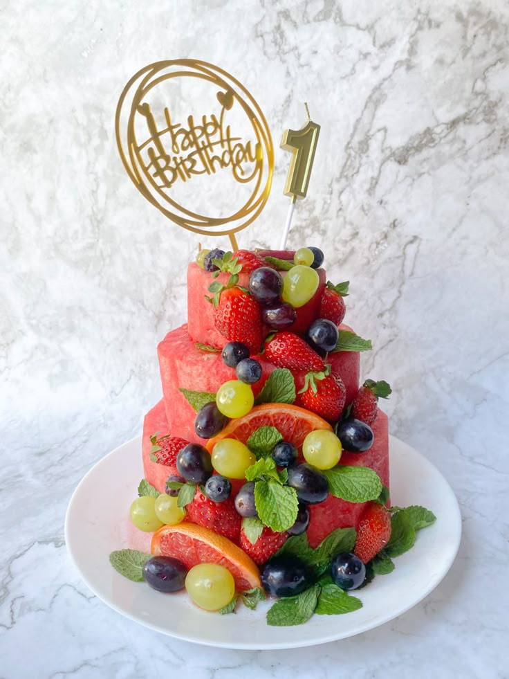 a watermelon cake with grapes and strawberries on top is decorated with a happy birthday candle