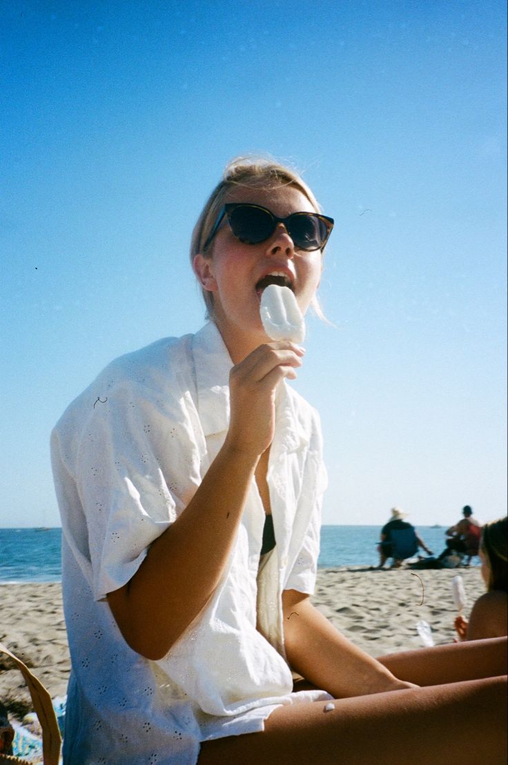 a woman sitting on top of a beach while holding a white object in her mouth