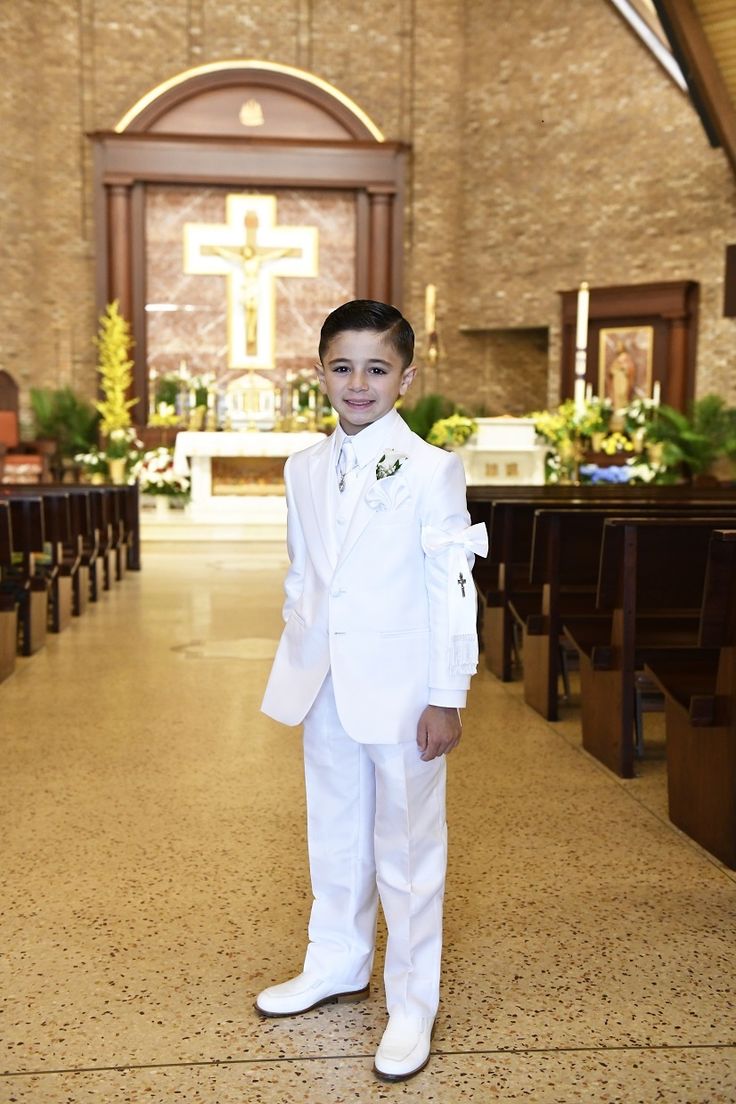 a young boy wearing a white suit and bow tie standing in front of pews