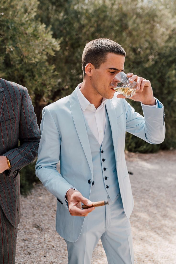 a man in a blue suit drinking from a glass next to another man wearing a white shirt and tie