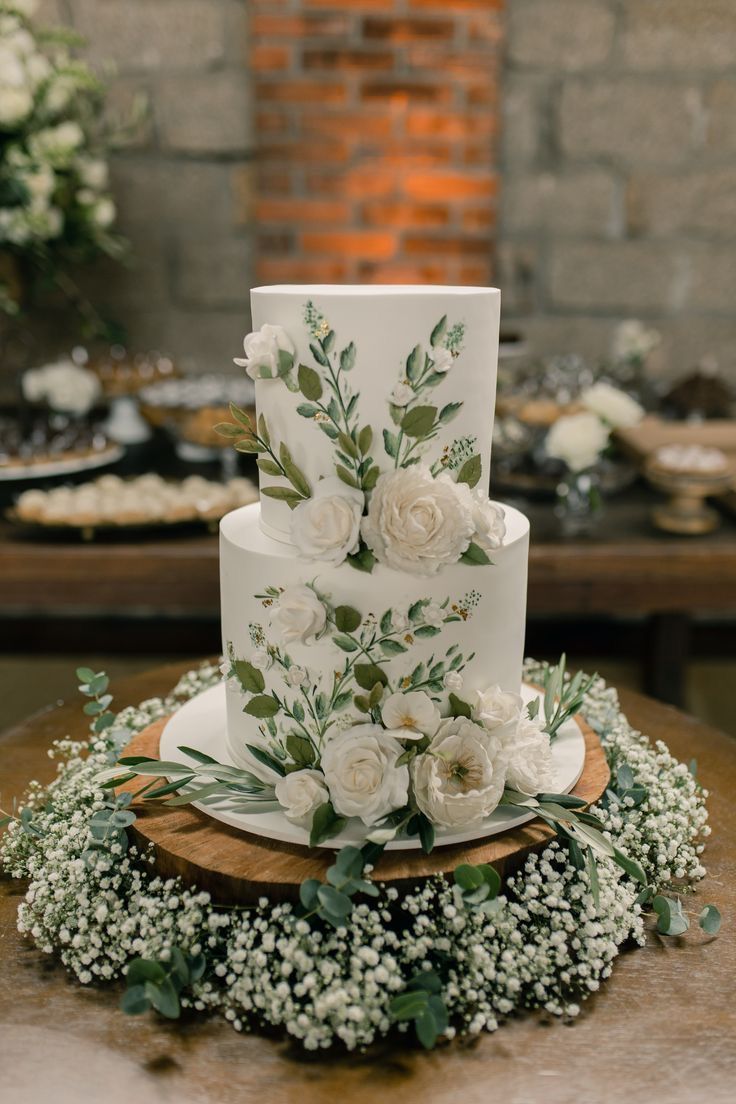 a wedding cake with white flowers and greenery sits on a table in front of a brick wall