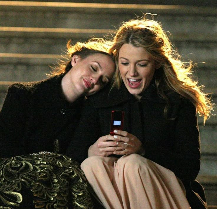 two women sitting on the steps looking at a cell phone and laughing with each other