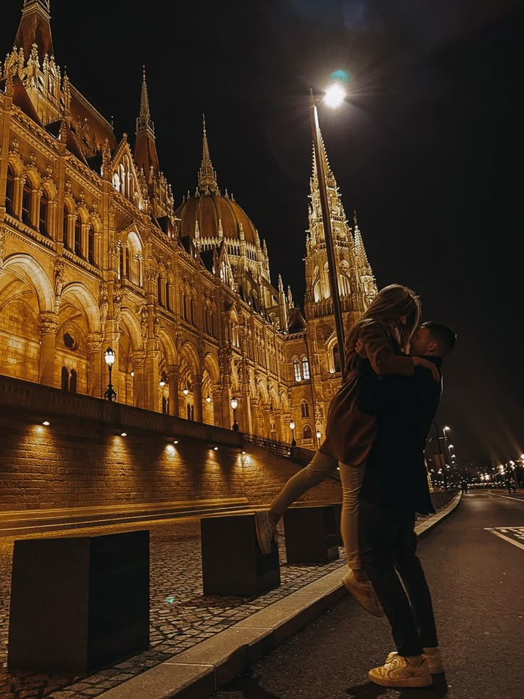 a man and woman kissing in front of a large building at night with lights on