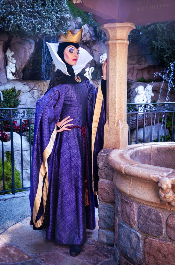 a woman wearing a purple dress and crown standing next to a fountain in front of a building