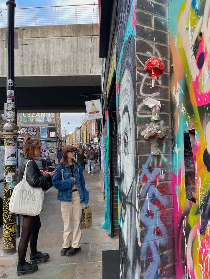 two women standing in front of a wall with graffiti on it and under a bridge