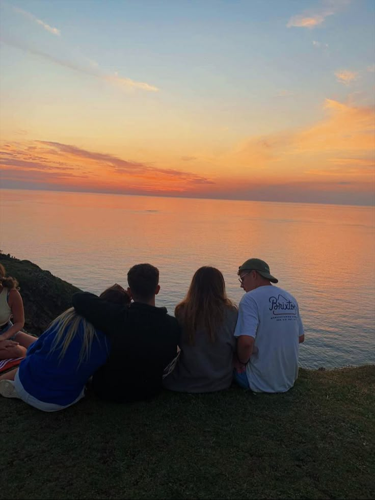 four people sitting on the edge of a cliff watching the sun set over the ocean