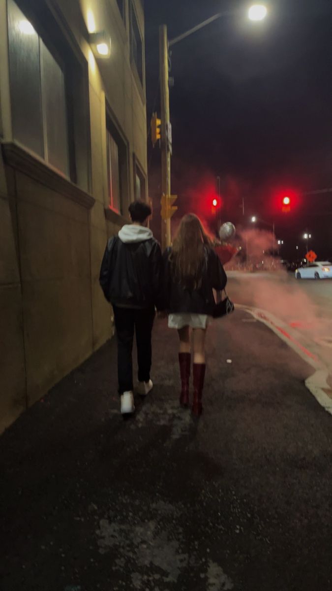 two people are walking down the street at night with red traffic lights in the background