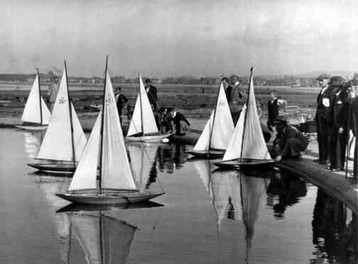 black and white photograph of sailboats on water with people standing around them in the background