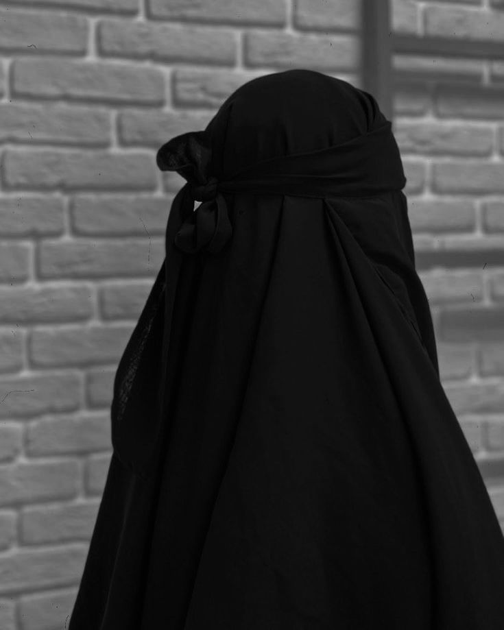 a woman wearing a black hijab standing in front of a brick wall with her back to the camera