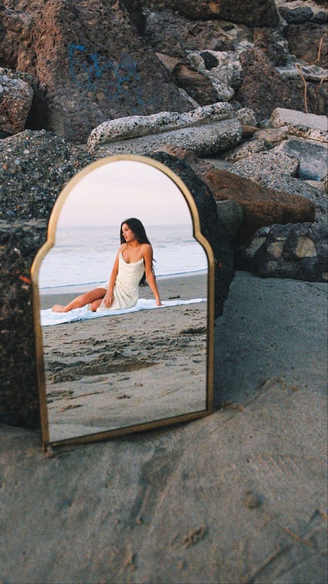 a woman sitting on the beach in front of a mirror