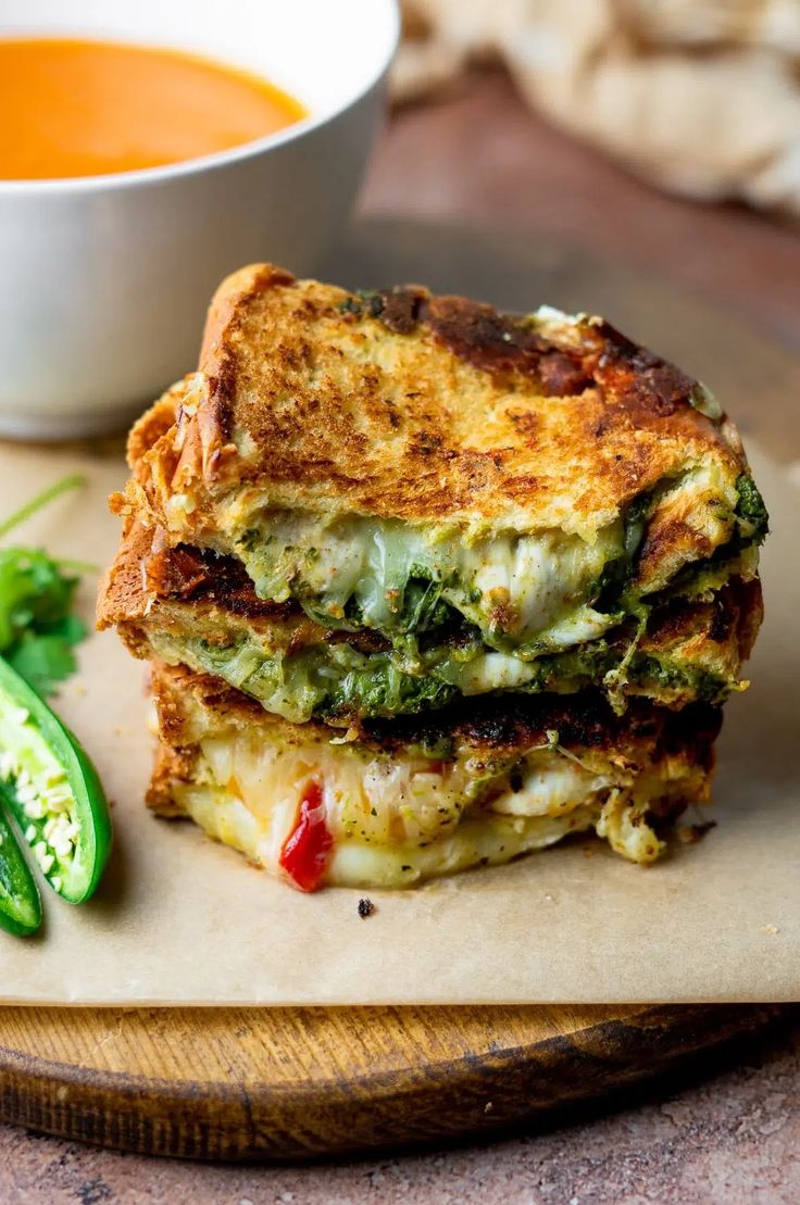 a grilled cheese sandwich on a cutting board next to a bowl of soup and some green peppers