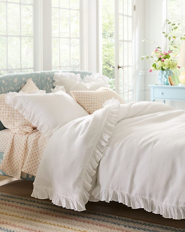 a bed with white sheets and ruffles in a room next to a window