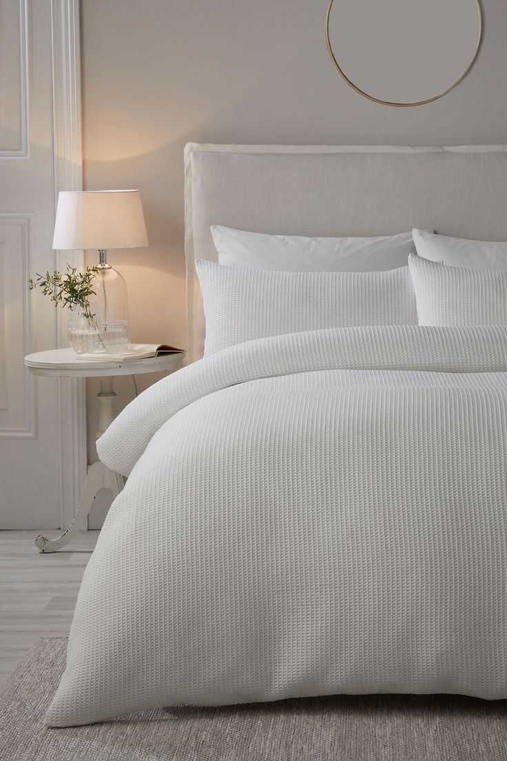 a white bed in a bedroom next to a lamp and table with a vase on it