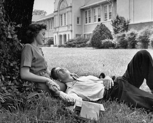 two women sitting on the grass in front of a house with a man laying down next to them