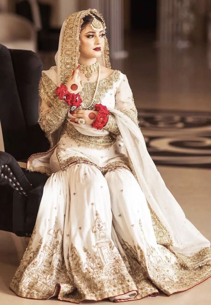 a woman in a white and gold wedding dress sitting on a black chair with red flowers