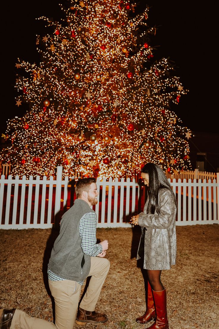 a man kneeling down next to a woman in front of a christmas tree