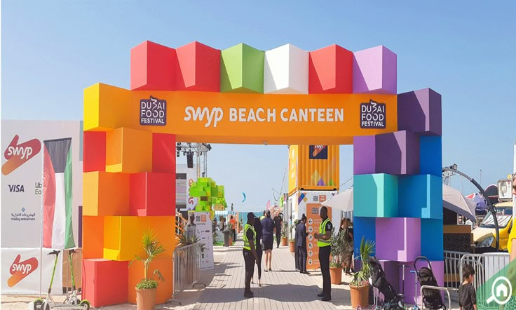 people are walking under an arch made out of colorful blocks and plants at the beach