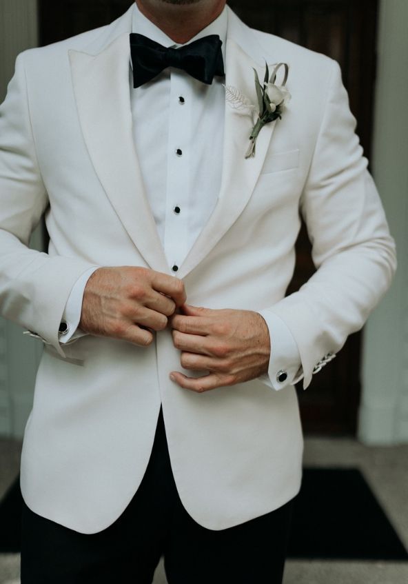 a man in a white tuxedo with his hands on his chest and wearing a black bow tie