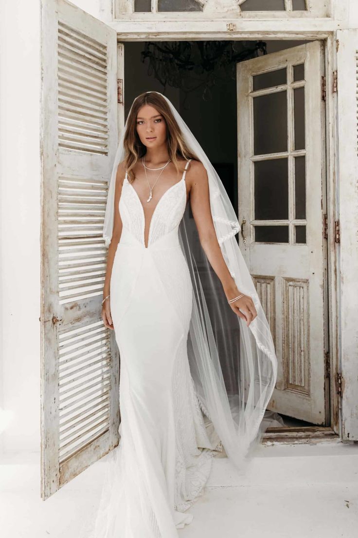 a woman wearing a wedding dress and veil standing in front of an open door with shutters