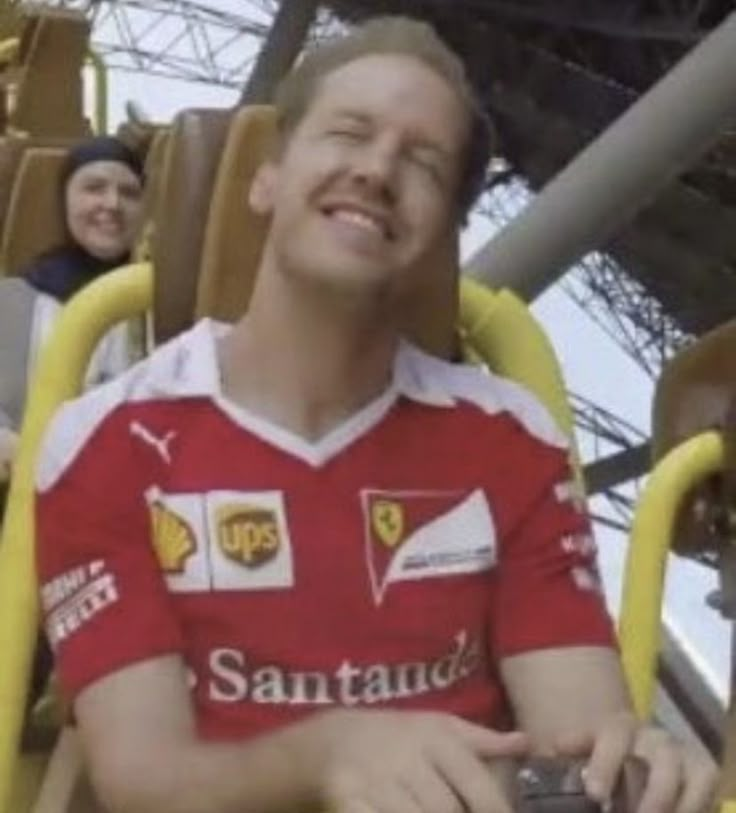 a man in a red shirt sitting on a yellow chair at an amusement park with other people behind him