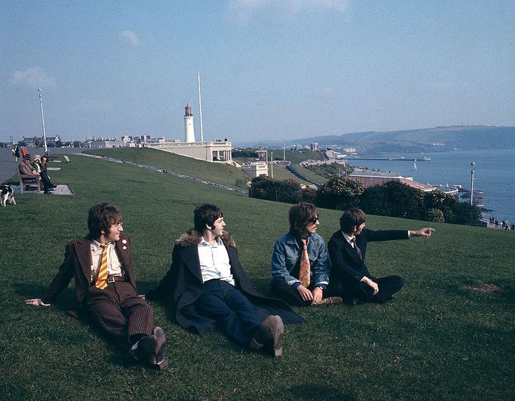 four young men sitting in the grass on top of a hill next to the ocean