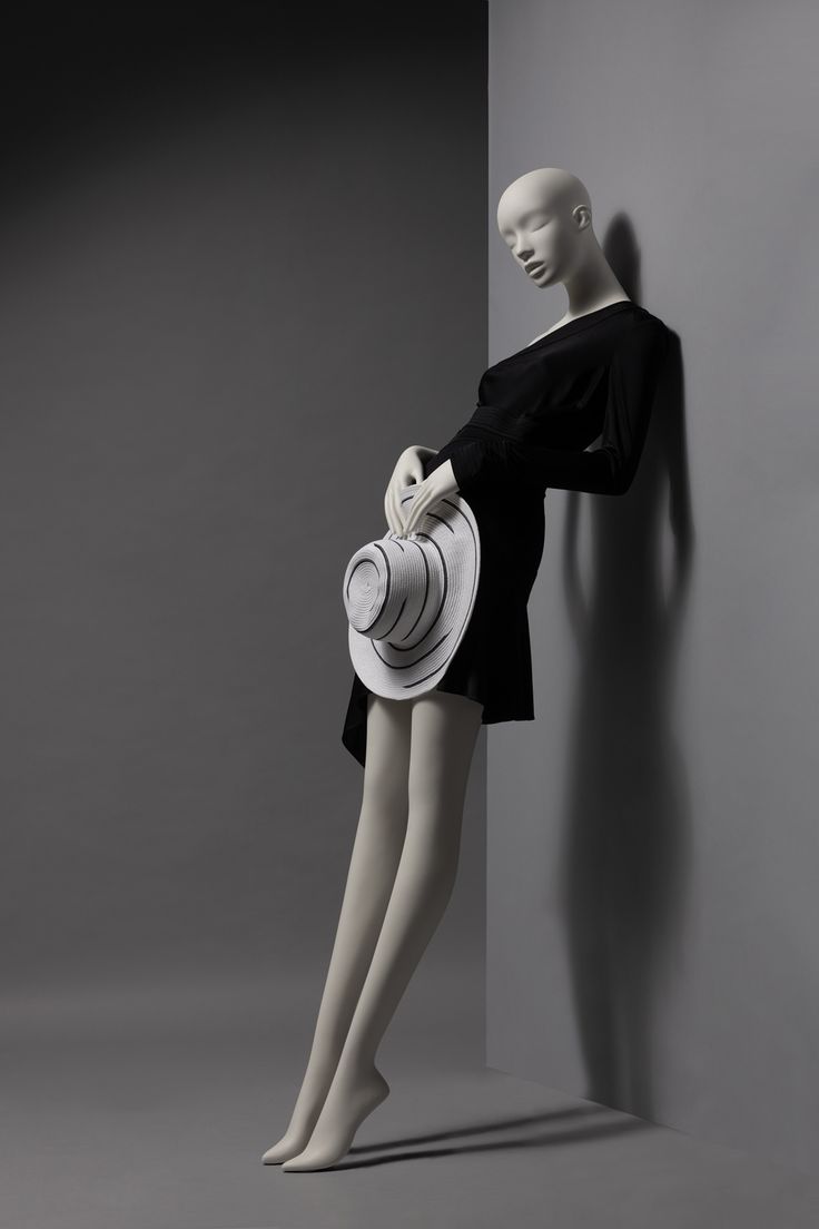 a mannequin wearing a black dress and white hat leaning against a gray wall