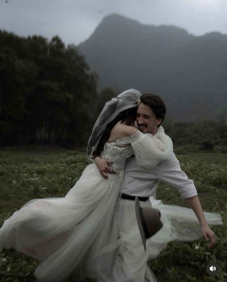 a man and woman are hugging in the grass with mountains in the backgroud