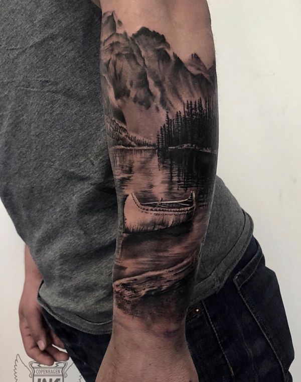 a man with a tattoo on his arm is standing in front of a lake and mountains