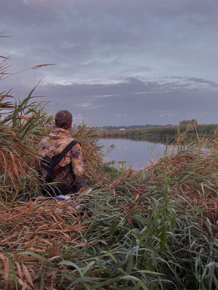 a person sitting on the edge of a body of water surrounded by tall grass and reeds