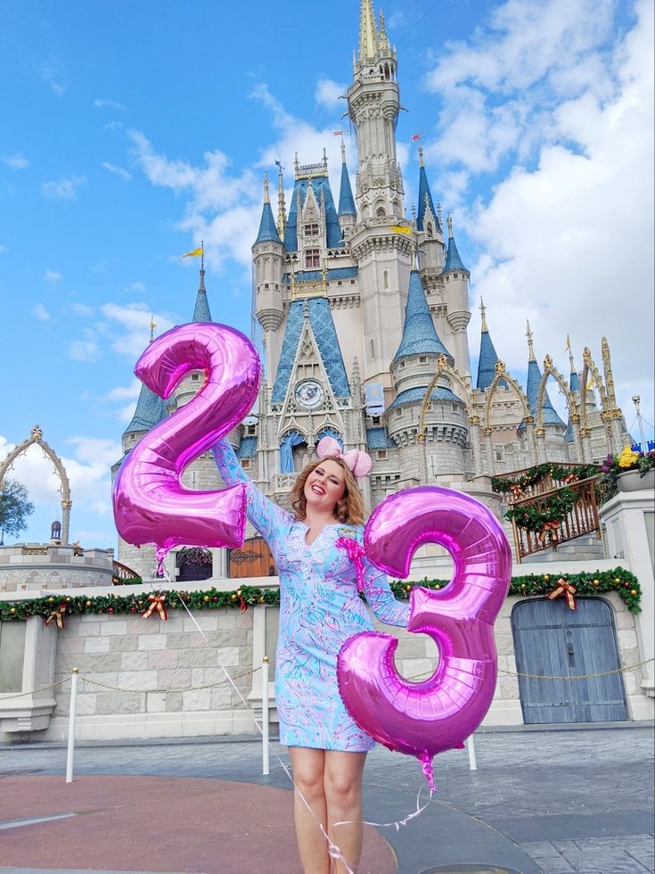 a woman in a blue dress is holding a pink number balloon with a castle in the background