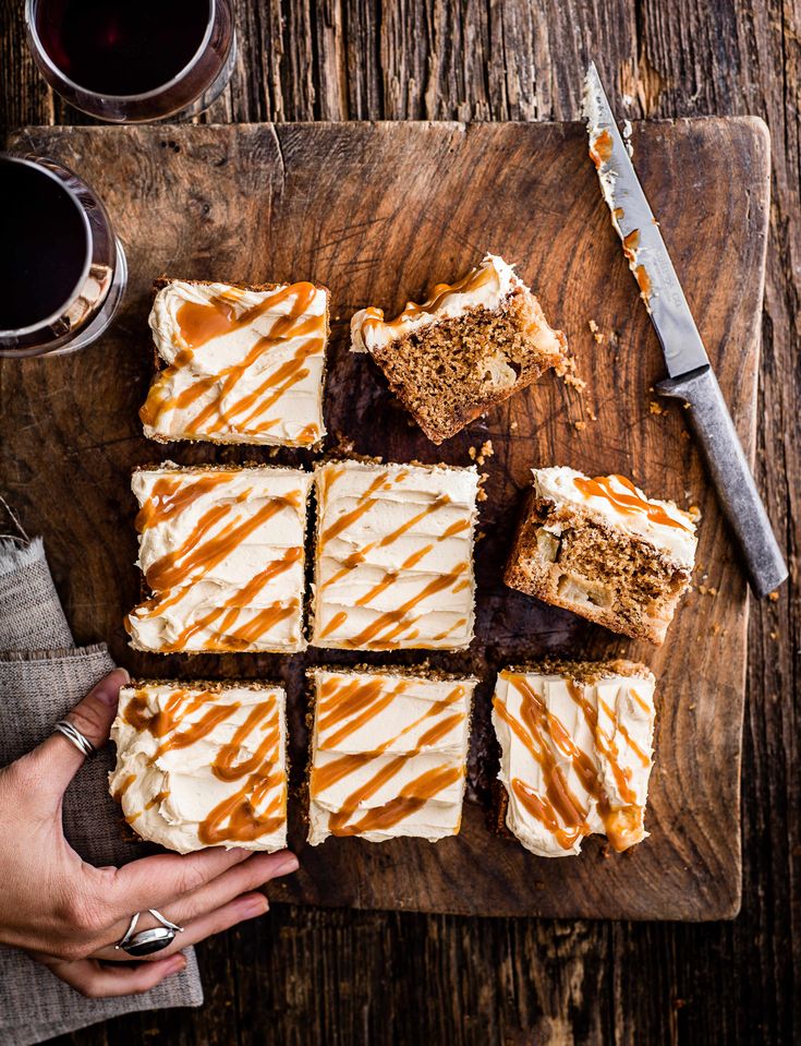 a person holding a knife over some desserts on a cutting board next to two cups of coffee