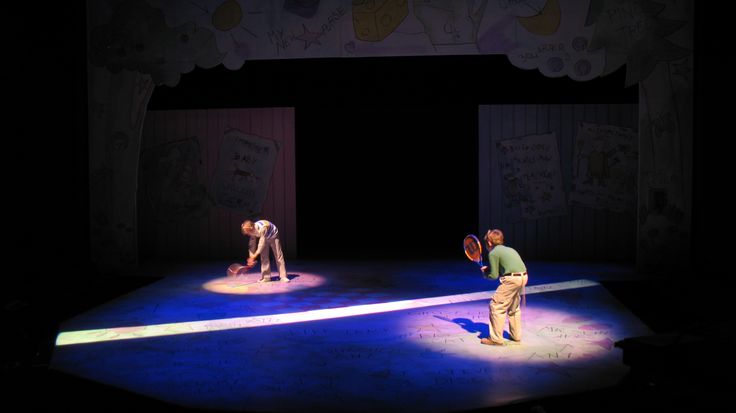 two people standing on a stage in the middle of a dark room, one holding a tennis racquet