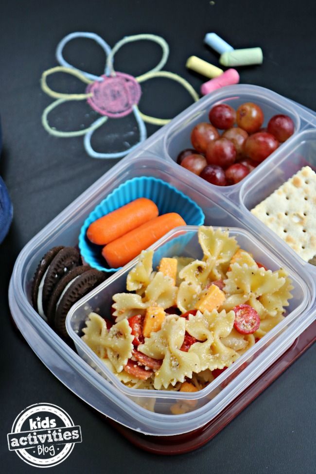 a plastic container filled with pasta and vegetables next to crackers, carrots and grapes