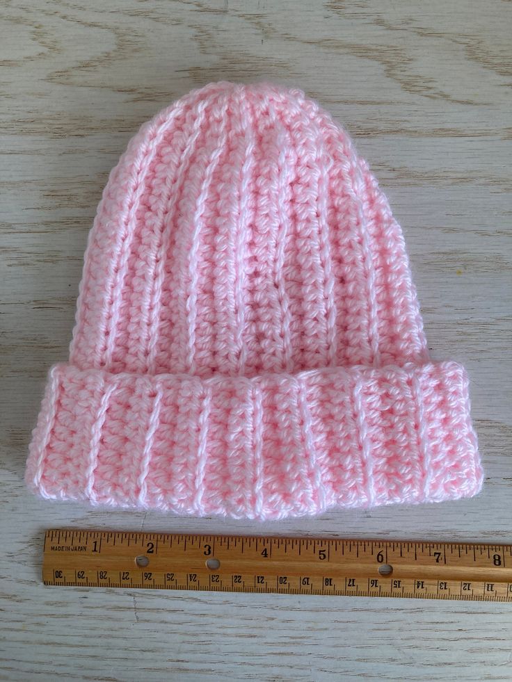 a pink knitted hat next to a ruler on a wooden table with a piece of tape