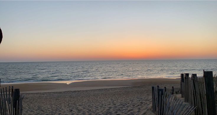 the sun is setting over the ocean and beach with a fence in front of it