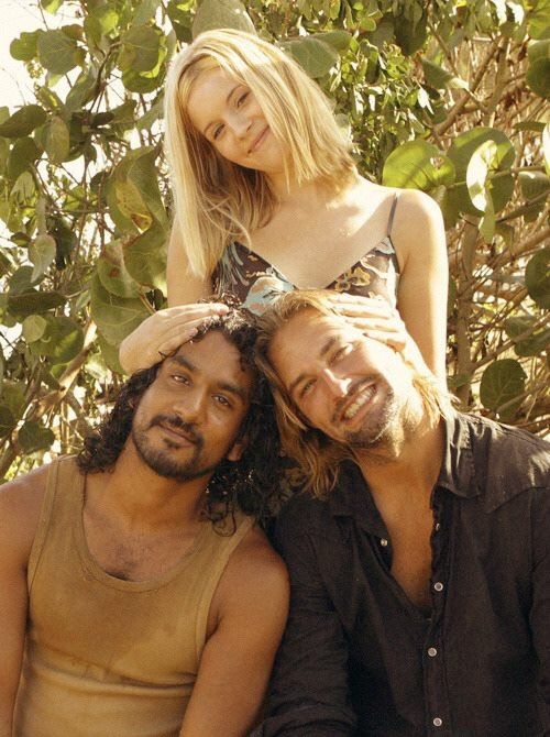 two men sitting next to each other in front of some trees and one woman standing behind them