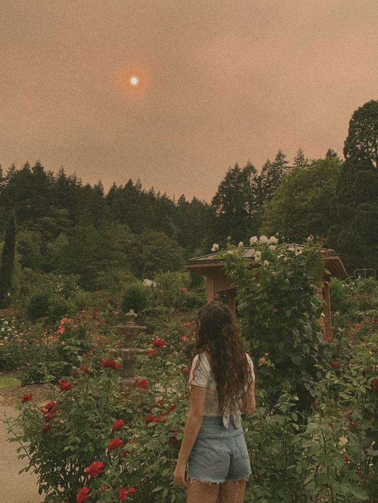 a woman standing in the middle of a garden with flowers and trees behind her, looking at the sun