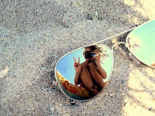 a woman taking a selfie in the reflection of her sunglasses on the beach,