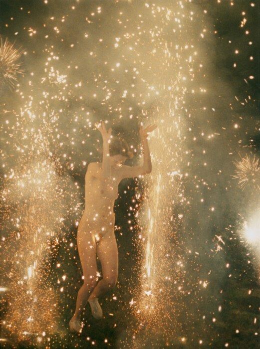 a naked woman standing in front of fireworks with her arms up to the sky and hands behind her back