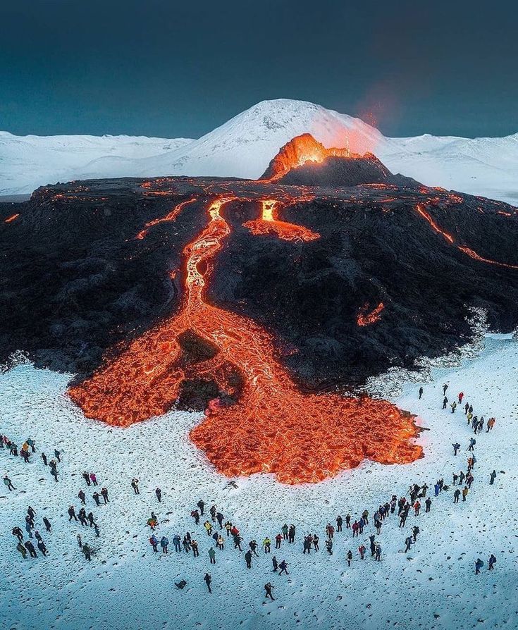 an aerial view of people standing in the snow near a mountain with lava pouring out of it