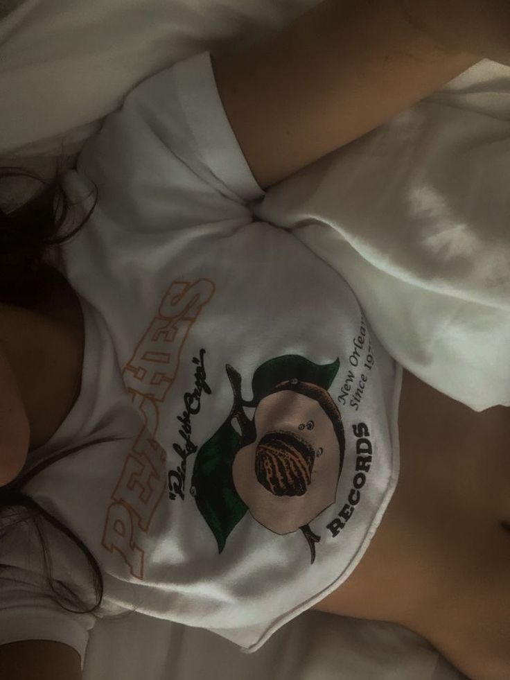 a woman laying on top of a bed wearing a white shirt with an orange design