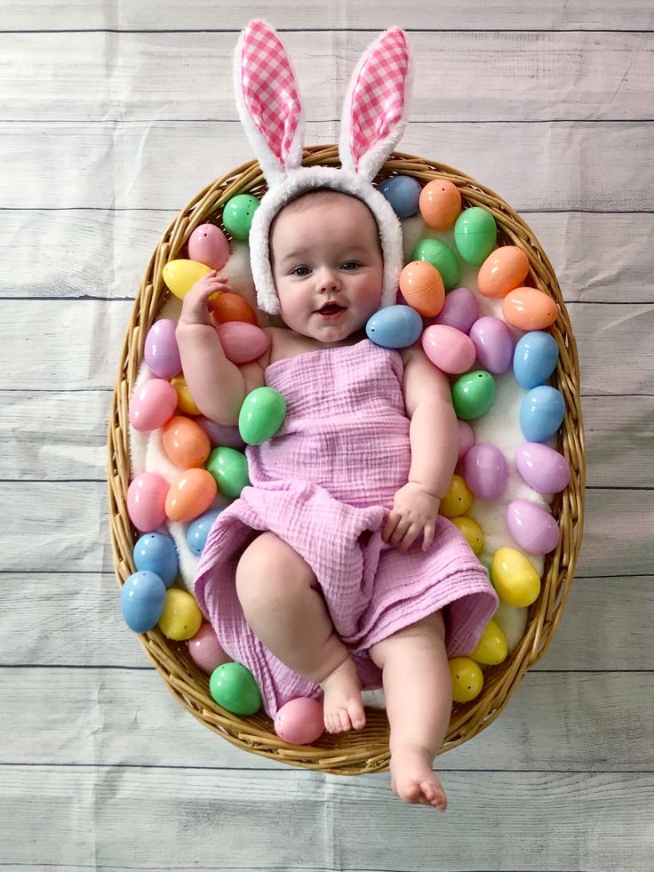 a baby wearing bunny ears laying in a basket filled with candy eggs and candies