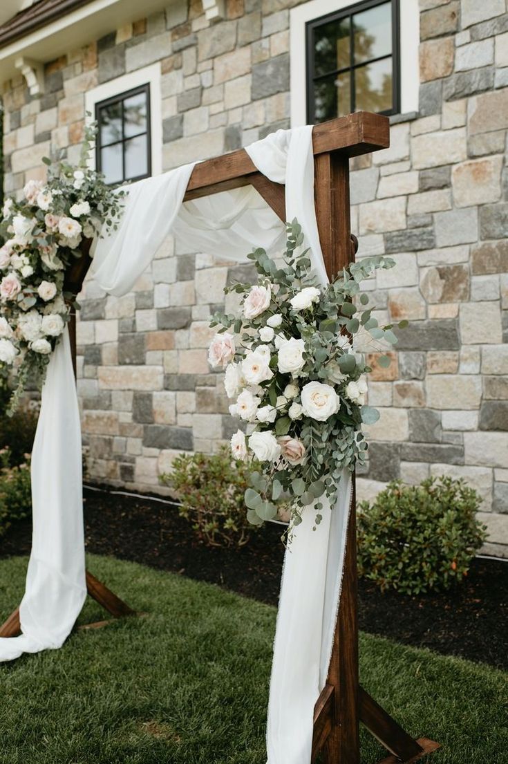 a wooden cross decorated with white flowers and greenery for an outdoor wedding ceremony in front of a stone building