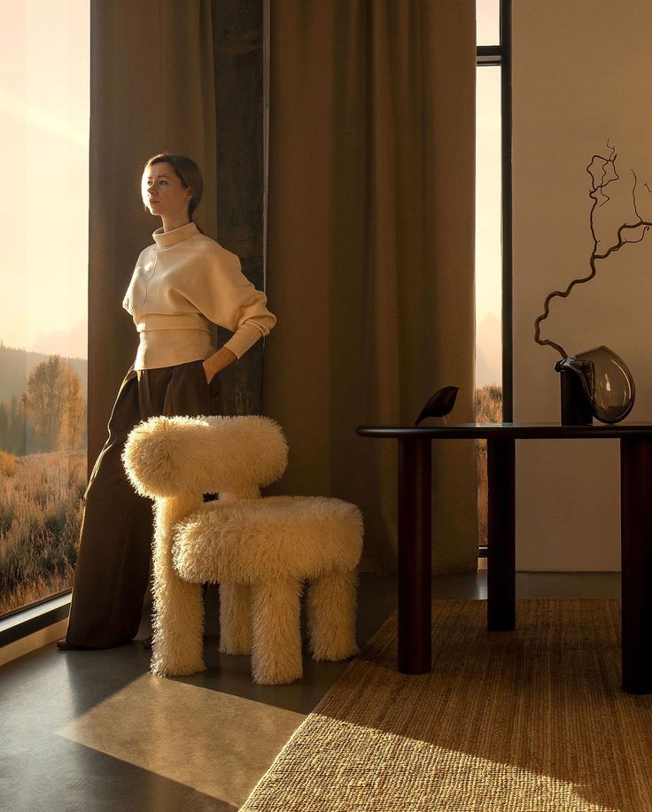 a woman standing in front of a window next to a chair and table with a dog on it