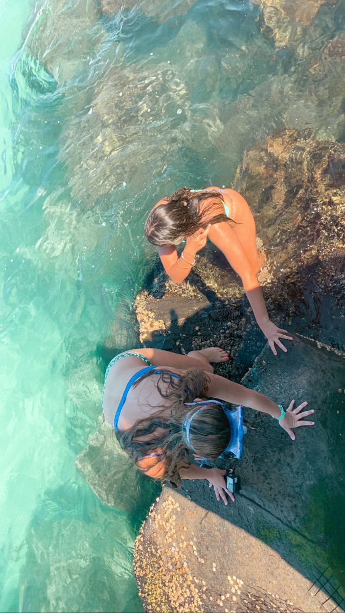 two women are standing in the water near some rocks and one is reaching for something