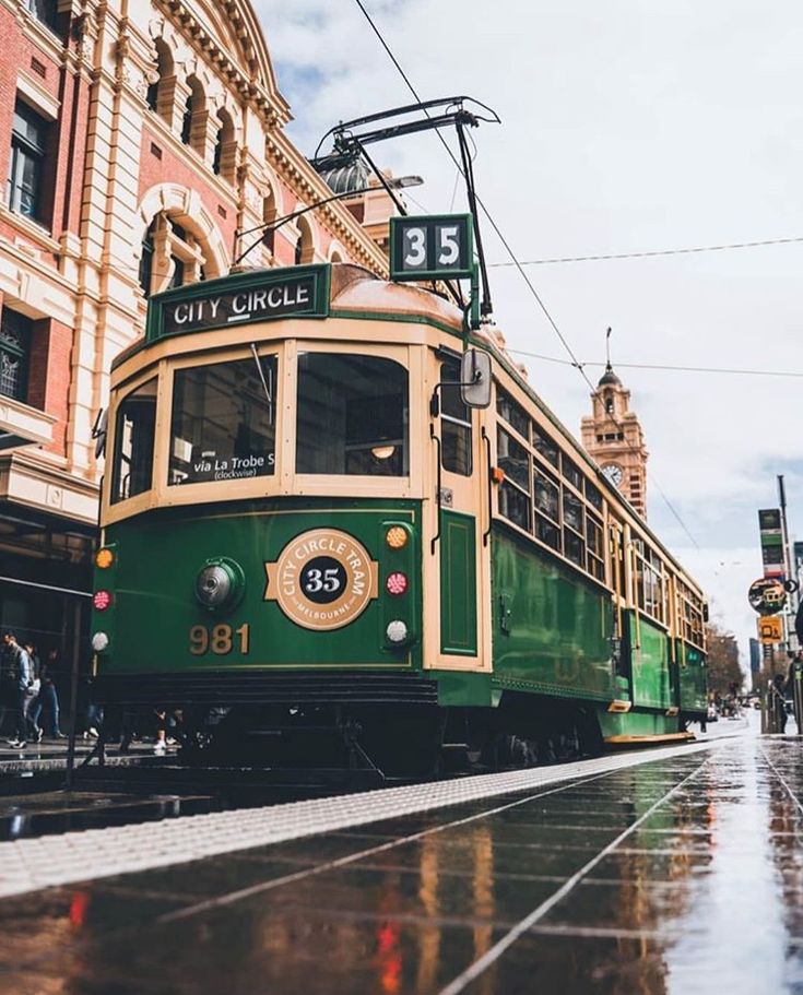 a green and white trolley car on street next to building with people walking by in the rain