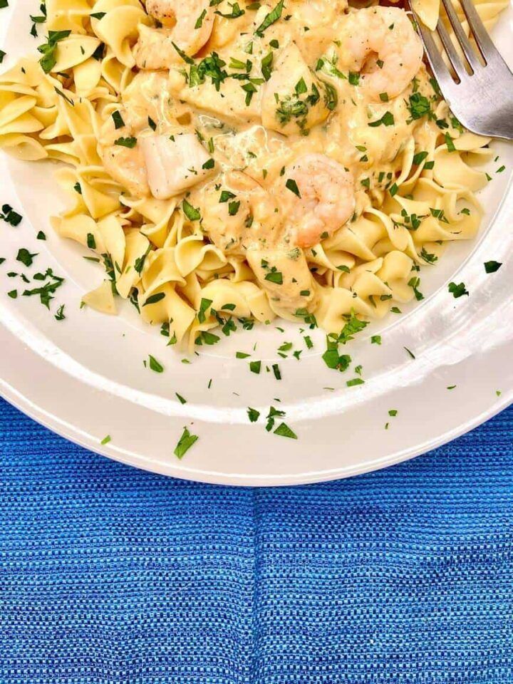 a white plate topped with pasta covered in sauce and shrimp next to a fork on a blue table cloth